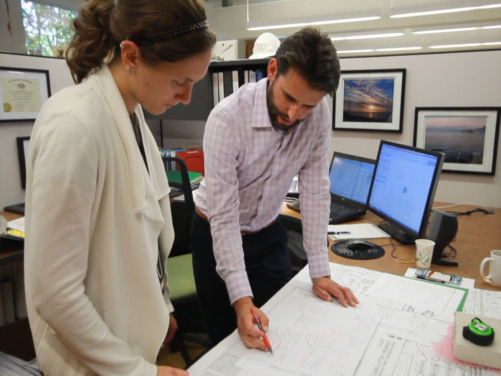 Peter Kerekgyarto, RA and Principal at Beers + Hoffman Architecture pointing to a plan on his desk and discussing with Sarah Faus CAD Designer